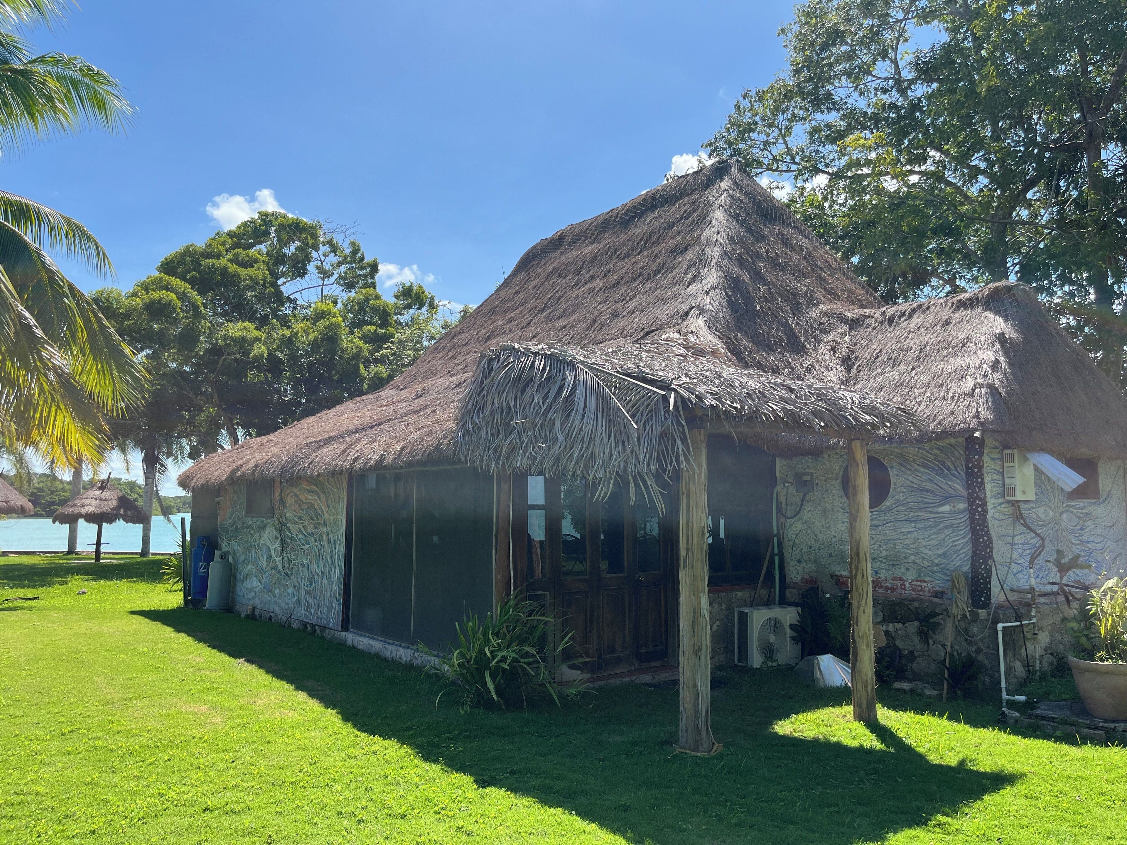A thatched-roof house with a covered porch, surrounded by lush greenery and a clear blue sky.