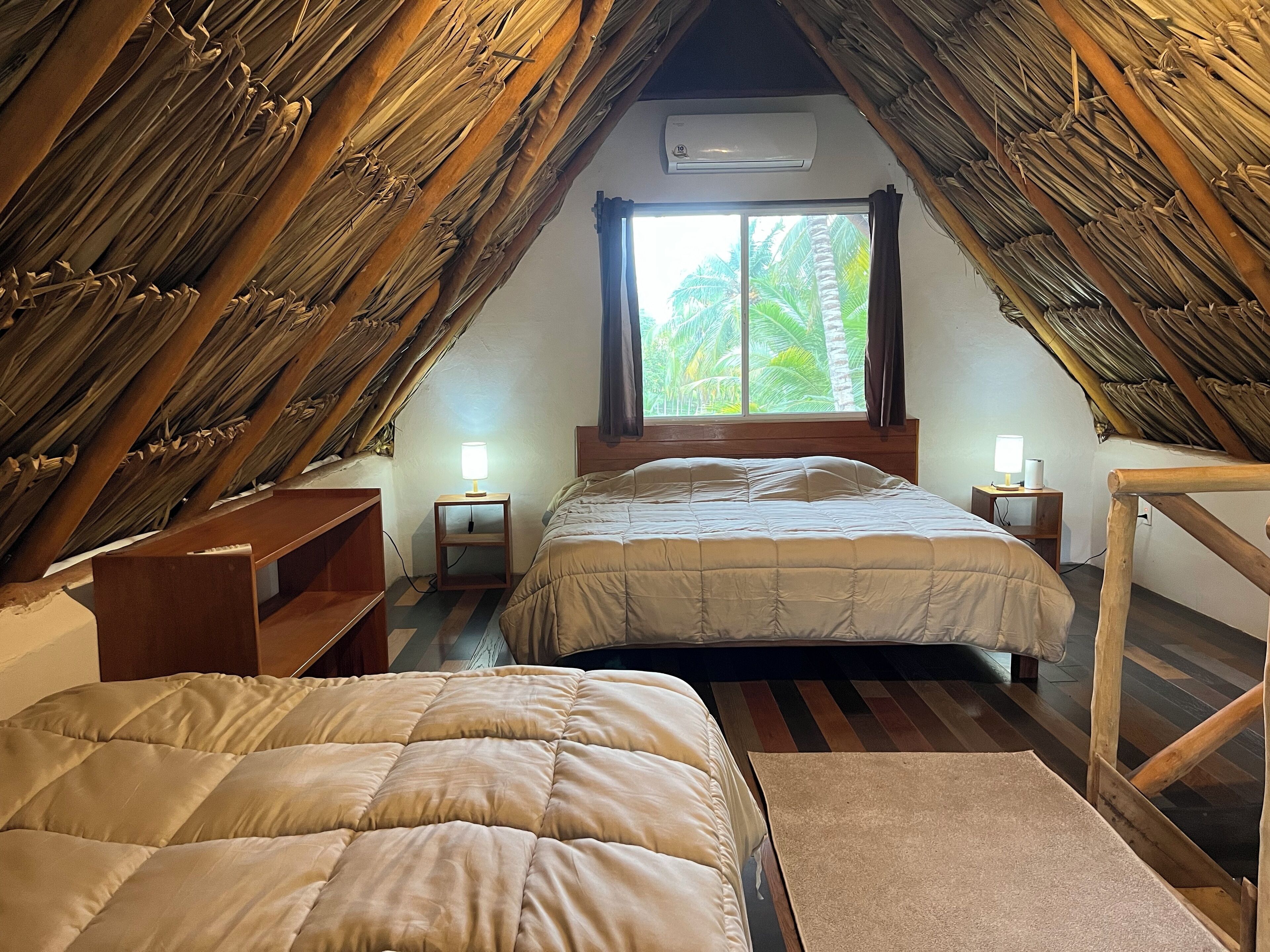 Quarto de sótão aconchegante, com teto inclinado, vigas de madeira e uma janela grande que oferece vista para palmeiras. Há duas camas com roupa de cama combinando e mesas de cabeceira com abajures.