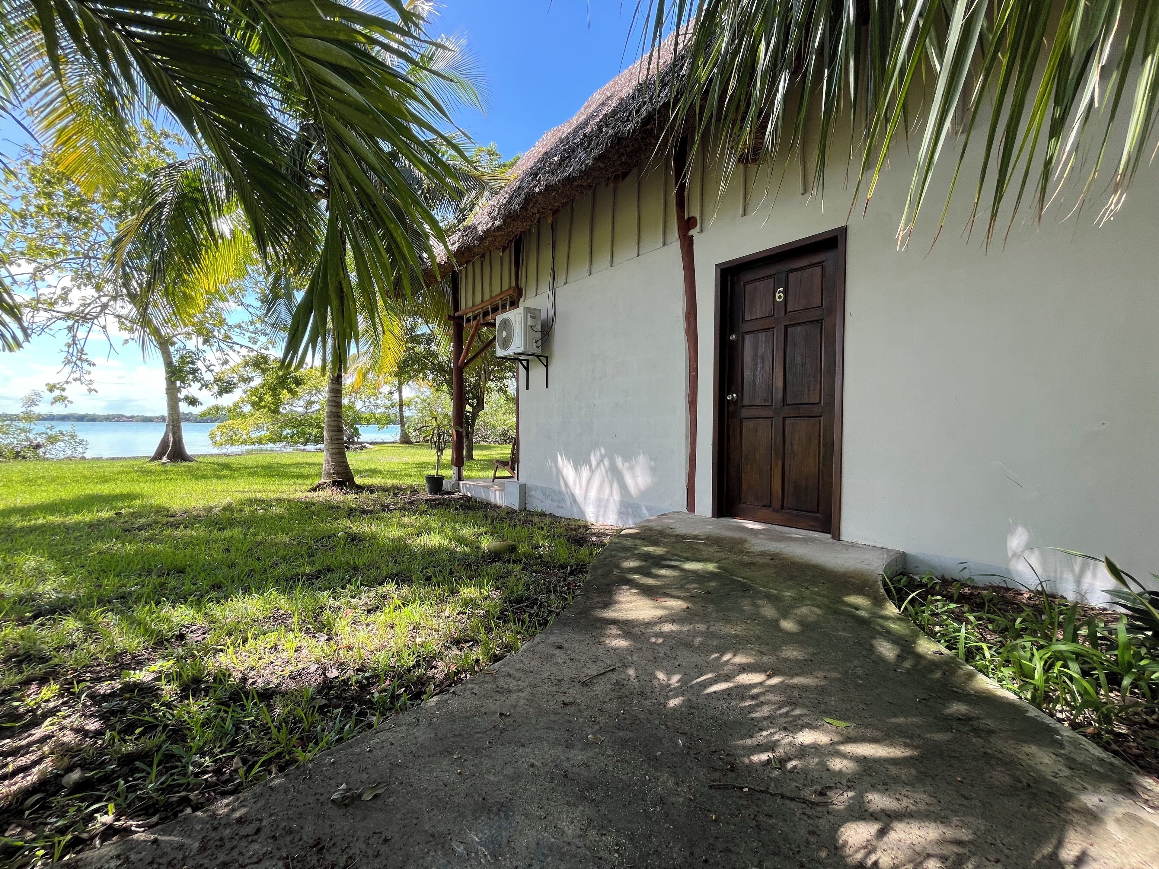 Uma casa com telhado de palha, um caminho de concreto e vista para um corpo d'água.