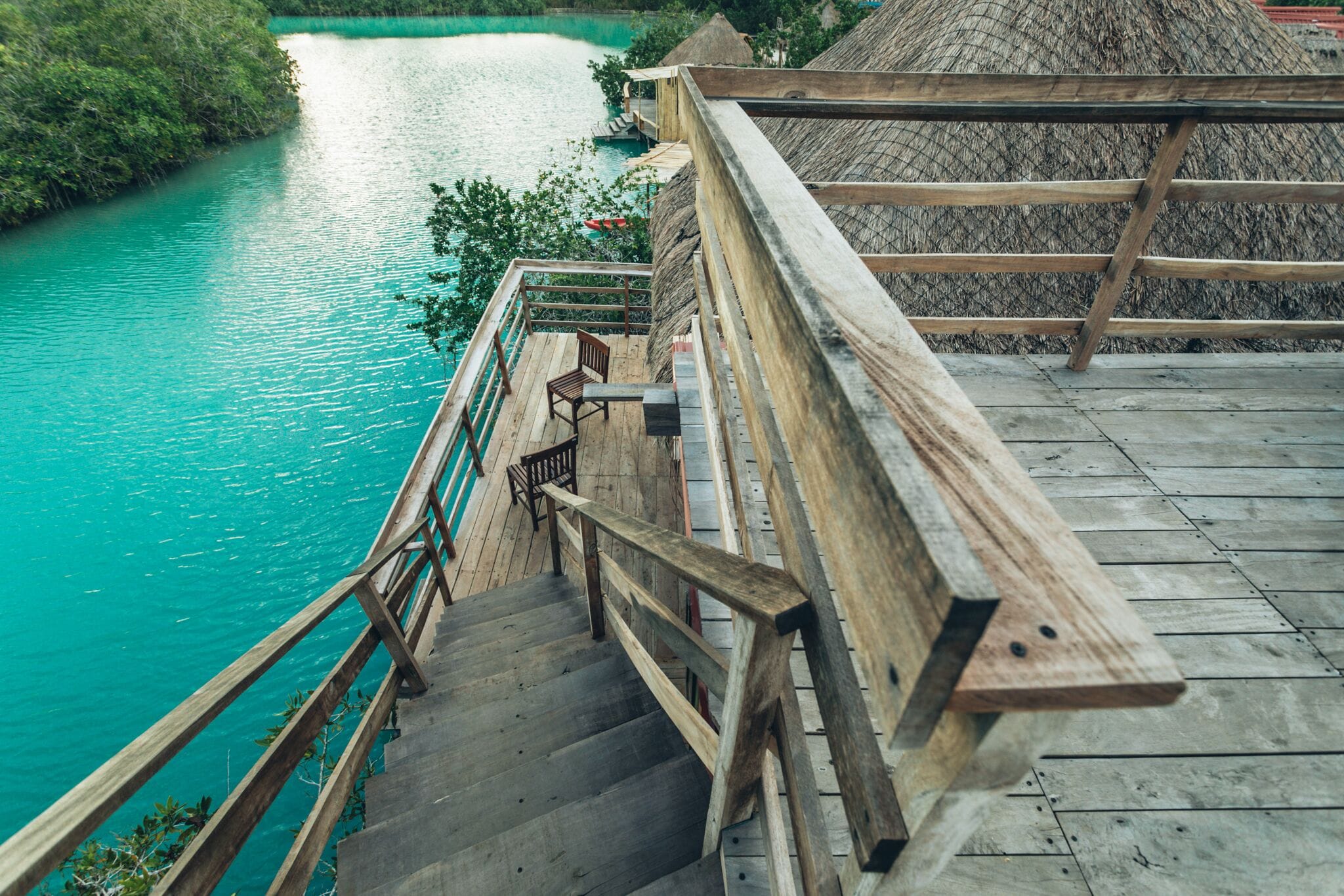 A wooden staircase leading down to a turquoise river, surrounded by lush greenery and a thatched-roof structure.