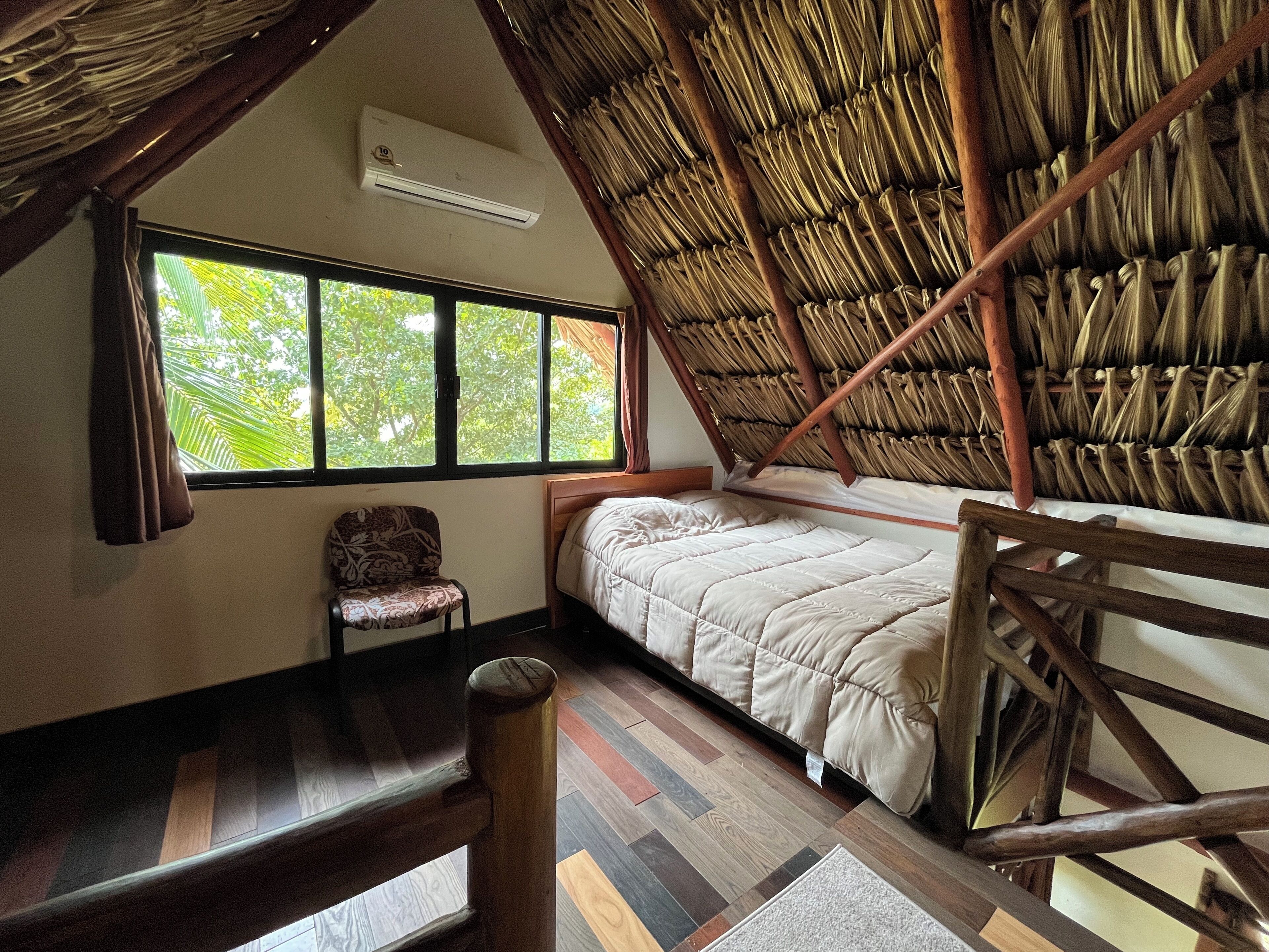 A room with a sloped ceiling, a bed, a chair, a window, and a wooden floor.