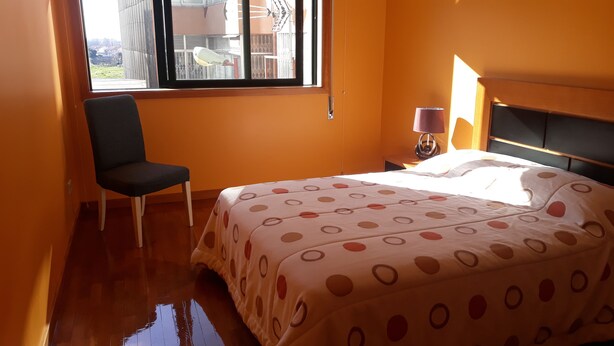 A bedroom with a bed, a chair, a nightstand, a lamp, and a window with a view of buildings. - Guida's place apartment (Porto)