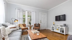 Un salon avec un canapé, une table basse et un téléviseur. Il y a une fenêtre avec des rideaux, un meuble blanc et une lampe.