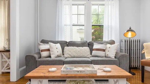 Un salon chaleureux avec un canapé gris, des coussins blancs, une table basse en bois et un radiateur.