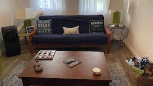 Een woonkamer met een donkerblauwe bank, twee kussens met tekst, een bijzettafeltje met een tijdschrift en een kleine schaal, en een lamp op een nachtkastje.