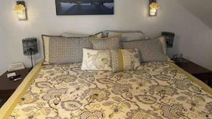 Een slaapkamer met een bed, beddengoed met een patroon, een nachtkastje, een lamp en een aan de muur hangend schilderij.