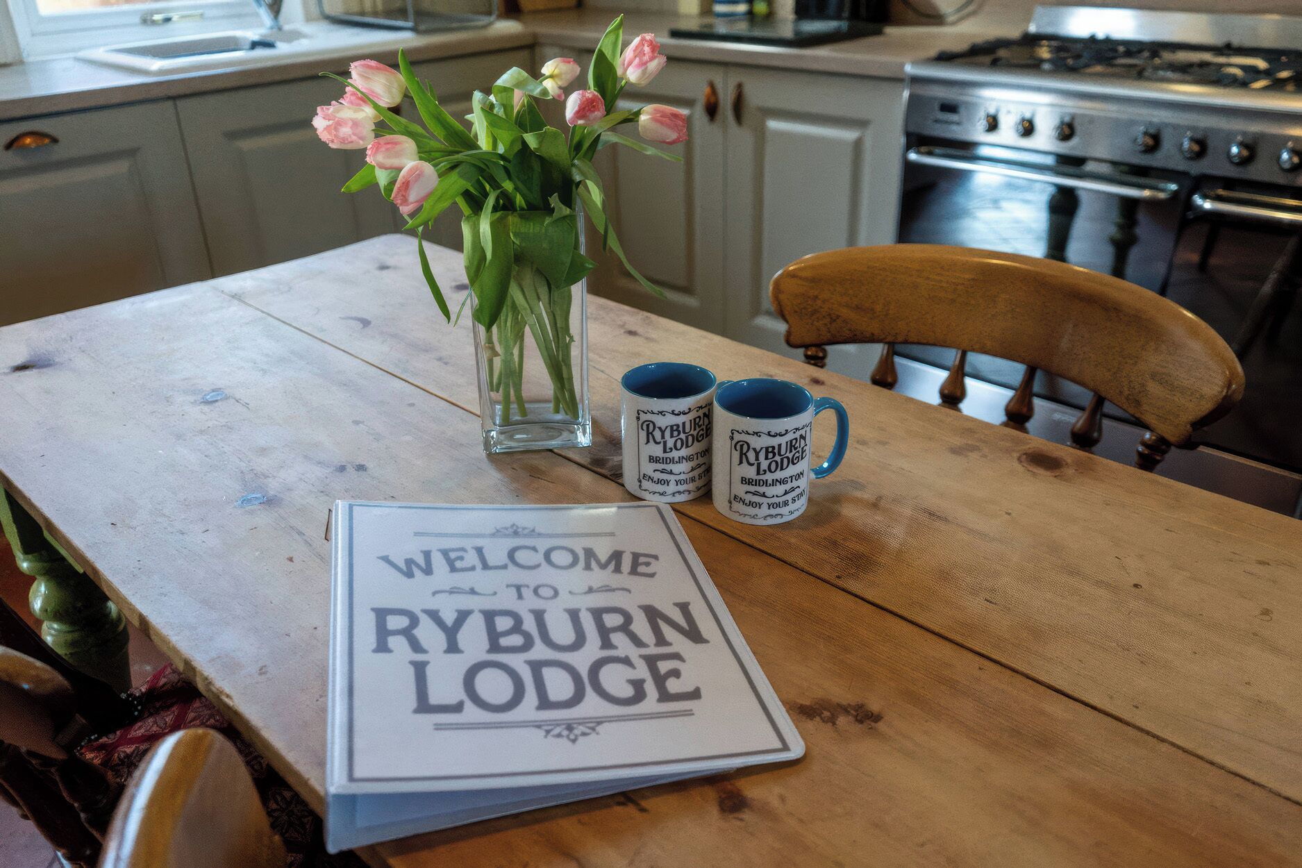 A wooden table with a welcome sign, two mugs, and a vase of flowers.