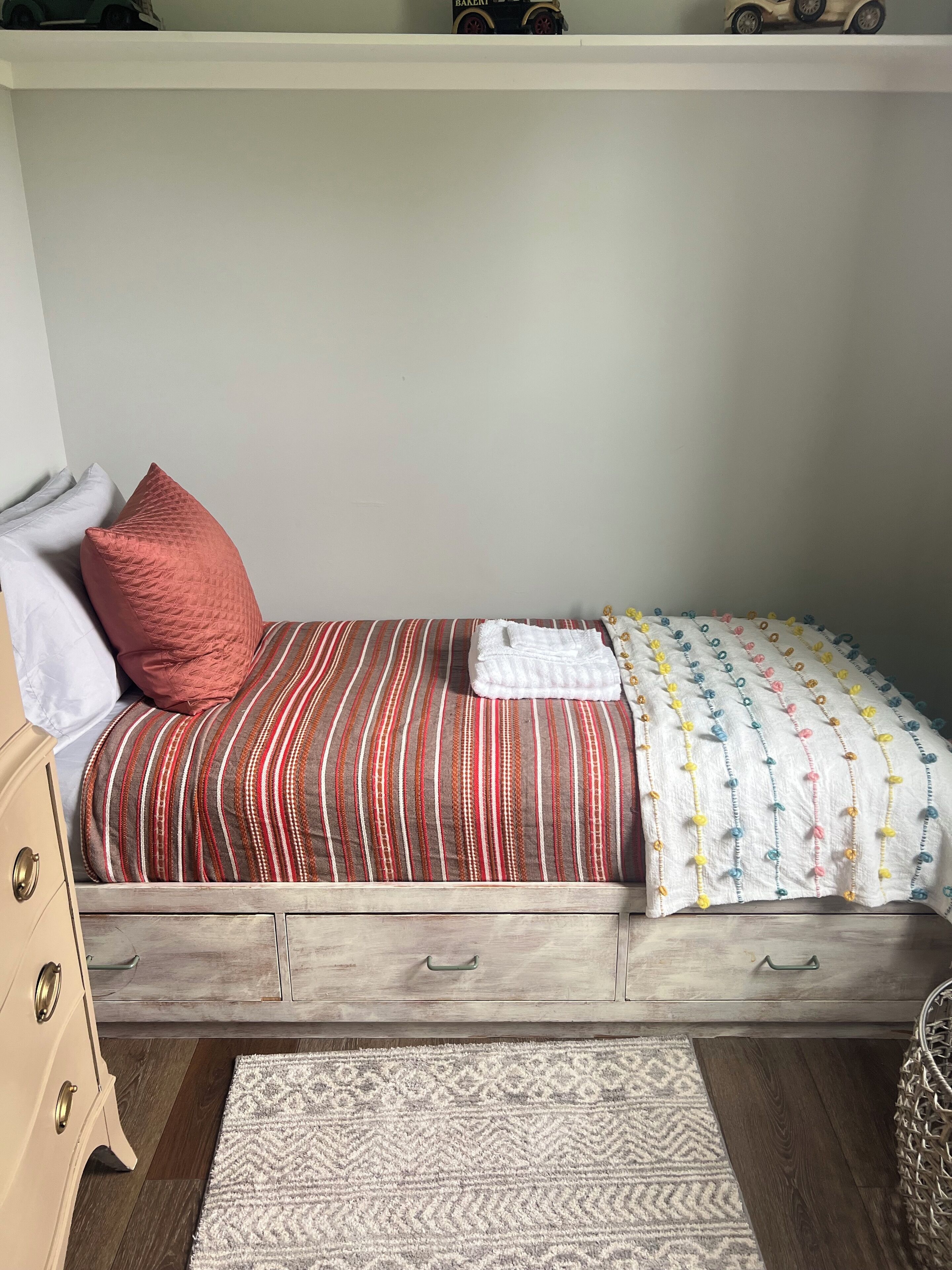 A single bed with striped bedding, a white pillow, and a red pillow. There is a white blanket with colorful pom-poms. A wooden nightstand with drawers is to the left.