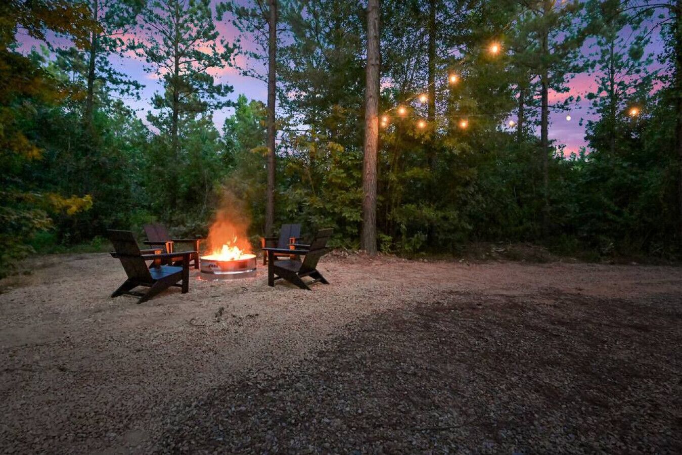 A campsite with two chairs around a central fire pit, surrounded by trees and string lights.
