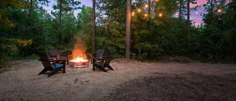A campsite with two chairs around a central fire pit, surrounded by trees and string lights.