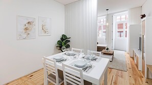 Ein moderner Essbereich mit einem weißen Tisch, gedeckt für vier Personen, einer Zimmerpflanze und Blick auf die Stadt durch große Fenster.