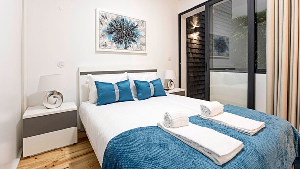 Ein modernes Schlafzimmer mit einem großen Bett, blauer Bettwäsche, einer Wanddekoration und Blick ins Freie durch eine Schiebetür.