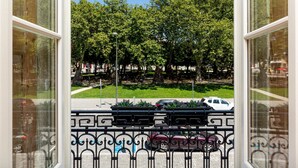 Une vue depuis une fenêtre, avec une rambarde en métal, un espace vert aménagé en parc et des voitures garées au loin.
