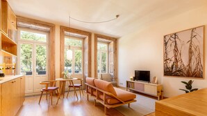 Un vaste espace de vie comprenant un canapé en bois, une table à manger avec des chaises et un téléviseur posé sur un meuble en bois. De grandes fenêtres laissent entrer la lumière naturelle et offrent une vue sur la verdure environnante.