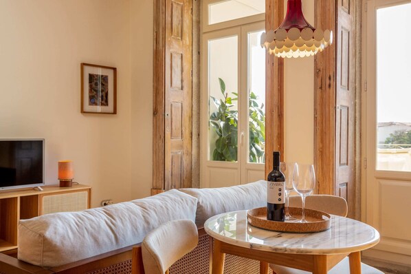 A cozy living room with a sofa, a small table with a bottle and glasses, a TV, and a window with a view of greenery.