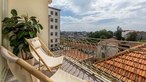 Een balkon met een stoel en een bloempot, met uitzicht op een stadsgezicht met terracotta daken.