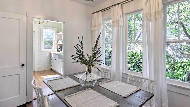 A dining area with a wooden table, white chairs, a vase with greenery, and large windows with white curtains.
