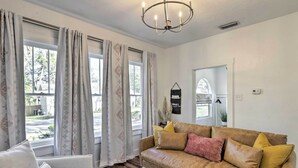 A living room with a beige sofa, a white armchair, and a patterned rug. There are large windows with curtains, a chandelier, and a wall-mounted mirror.