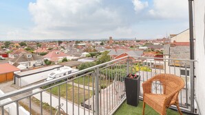A balcony with a wicker chair, a black planter with flowers, and a view of a residential area with houses and a tower in the distance. - Ty Uchaf (Rhyl)