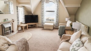 A cozy living room with a fireplace, a television, and a view of the beach. - Ty Uchaf (Rhyl)