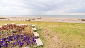 A coastal landscape with a grassy area, a flower bed with purple flowers, and a view of the beach and sea under a cloudy sky. - Ty Uchaf (Rhyl)