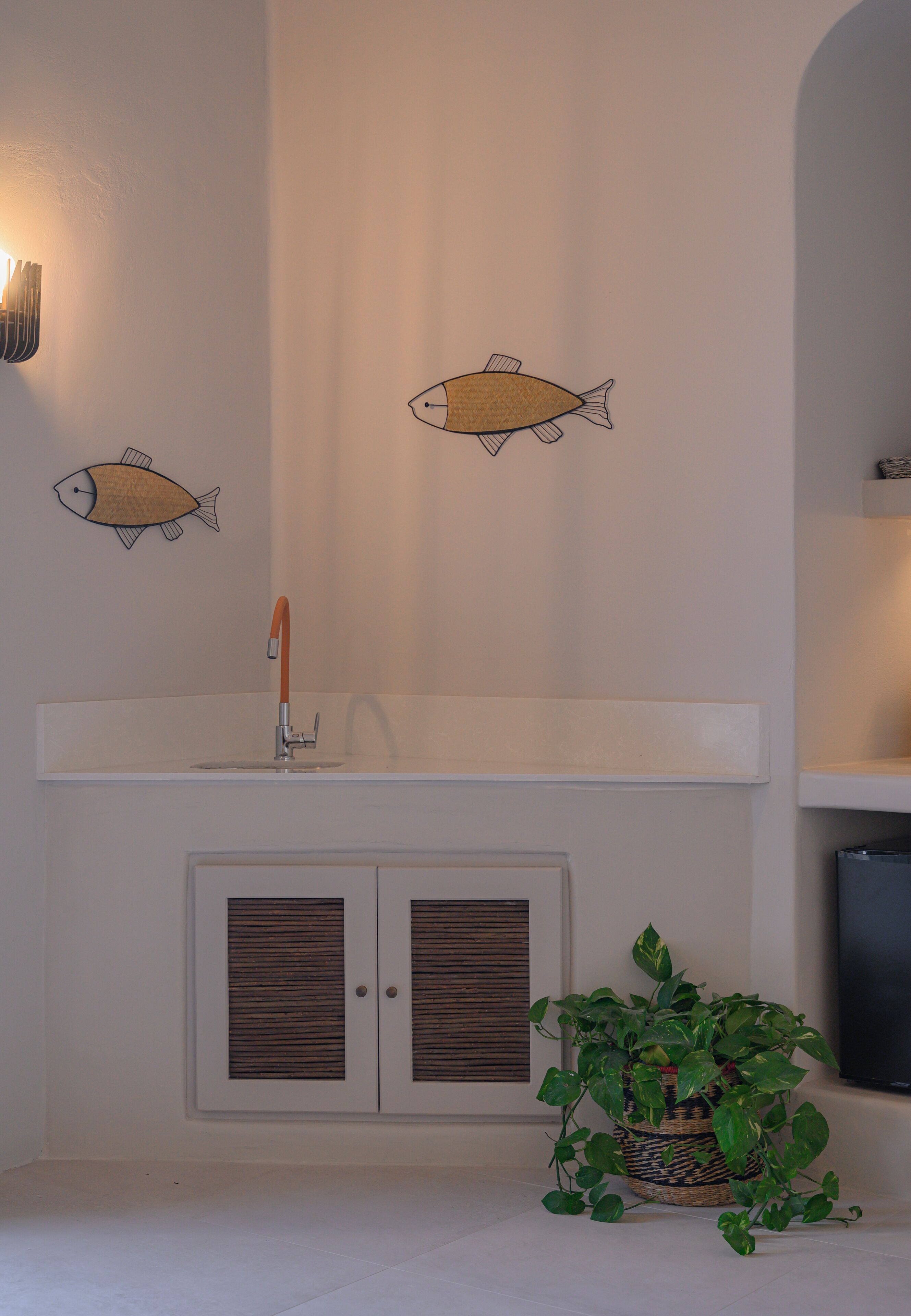 Et badeværelse med en hvid vask, to fiskemotiver på væggen, en potteplante og et fjernsyn på en hylde.