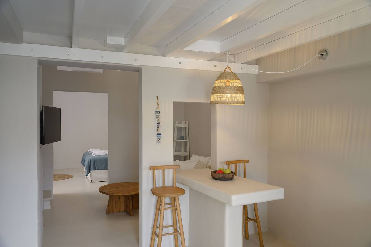 Una habitación moderna y minimalista con una paleta de colores blancos, que incluye una zona de comedor con taburetes de madera, una mesa blanca y una lámpara colgante de fibra natural.