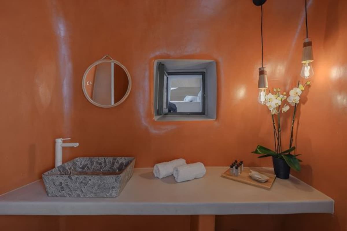 Une salle de bain avec un lavabo en marbre, un miroir rond, une petite fenêtre et une orchidée en pot.