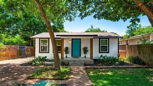 A single-story house with a front porch, a blue door, and a small front yard with a tree and plants. - Spacious Patio + Close to The Square + Pet Friendly + Full Kitchen (San Marcos)