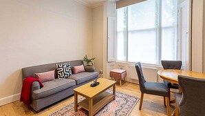 A living room with a grey sofa, a wooden coffee table, a round table with chairs, and a large window providing natural light.