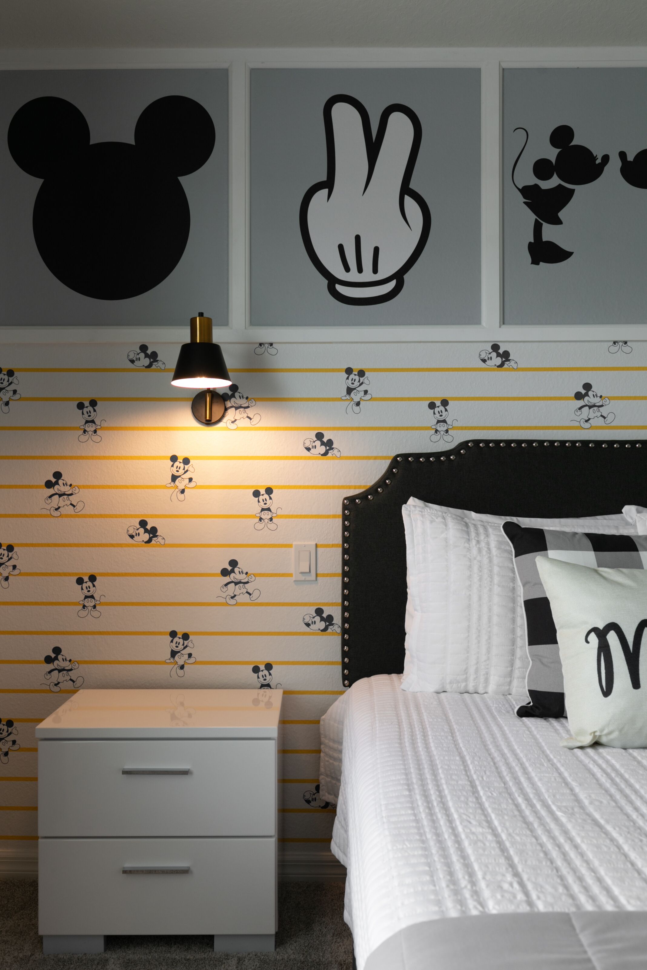 Quarto com papel de parede temático do Mickey Mouse, uma cama com roupa de cama branca, uma mesa de cabeceira e uma luminária fixada na parede.