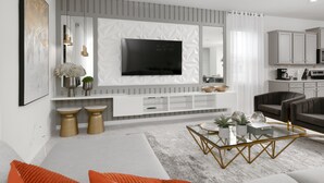 A modern living room with a flat-screen TV mounted on a textured wall, a glass coffee table, and a grey sofa with orange cushions.