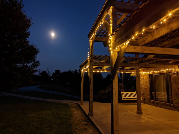 Una pérgola de madera con luces colgantes, un banco y un muro de piedra.