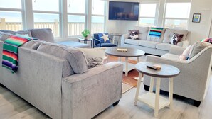 A spacious living room with a grey sofa, a blue armchair, and a grey armchair. There is a flat-screen TV mounted on the wall, a small round table, and a coffee table. The room has large windows with a view of the sea. - Beachfront Retreat l Hot Tub l Gulf Views, Wraparound Deck, Sleeps 12, Pet-Friendly, & More (Gilchrist)