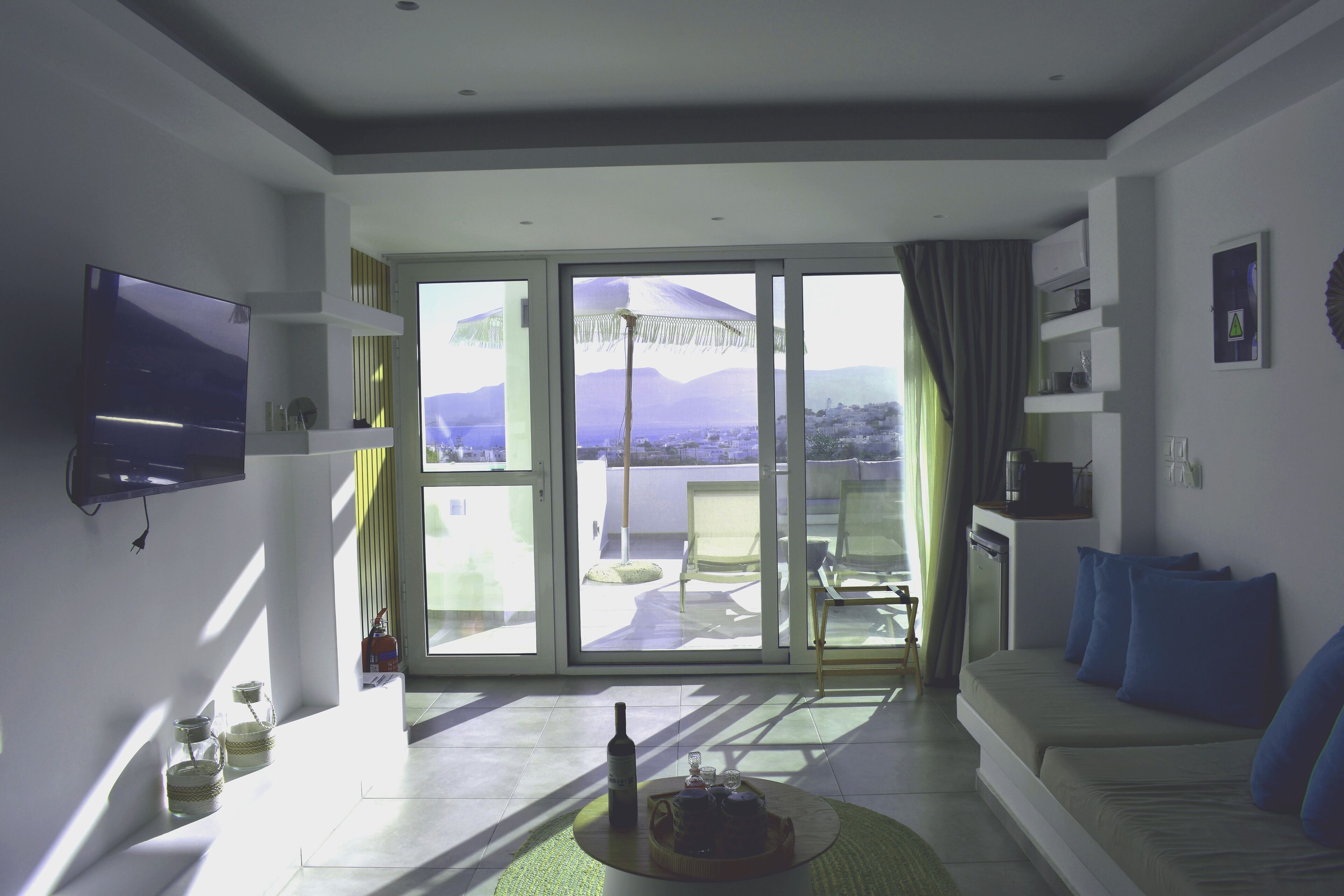 A modern living room with a flat-screen TV, a sofa with blue cushions, a coffee table with a bottle and glasses, and a large sliding glass door leading to an outdoor patio with a view of mountains.