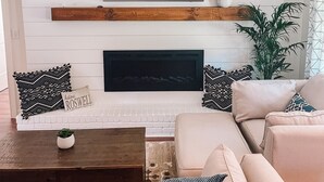 Un salon moderne avec un mur en briques blanches, un téléviseur à écran plat fixé au-dessus d’une cheminée, et un coin salon confortable avec un canapé beige et des coussins à motifs.