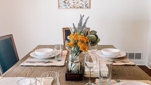 Une table à manger dressée avec de l’assiette blanche, des verres et des couverts, agrémentée d’un arrangement floral placé au centre, dans un vase.