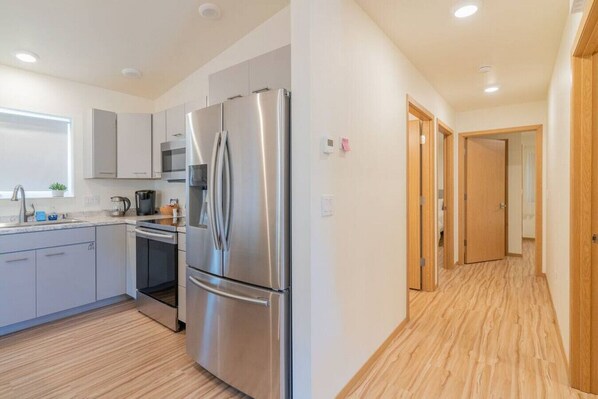 A modern kitchen with stainless steel appliances, a microwave, and a sink. There is a wooden floor and a hallway leading to another room. - The Northern Bungalow With Modern Charm (Fairbanks)