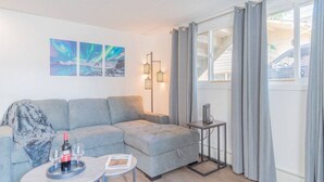 Een moderne woonkamer met een grijze modulaire bank, een klein rond tafeltje met twee wijnglazen en een fles, en een vloerlamp. Twee ingekaderde foto’s hangen aan de muur en er is een raam met grijze gordijnen.