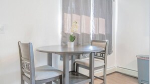 Een kleine, ronde eettafel met twee stoelen, een bloempot en een raam met dunne gordijnen.