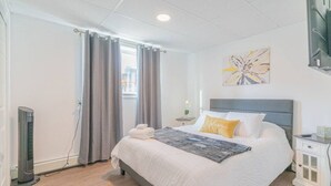 Een netjes opgemaakt slaapkamer met een bed, nachtkastje, een tv aan de muur en een raam met gordijnen.