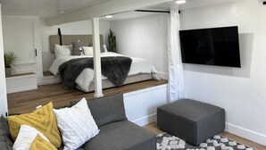 A modern bedroom with a bed, a grey sofa, a grey ottoman, and a TV mounted on the wall. - Mountain Views/Studio in Montecito. 7-10 min to beach or downtown SB (Montecito)