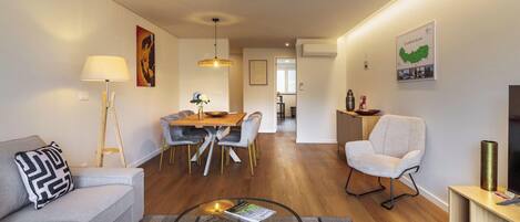 A modern living room with a sofa, a dining table with chairs, a glass coffee table, and a TV stand.