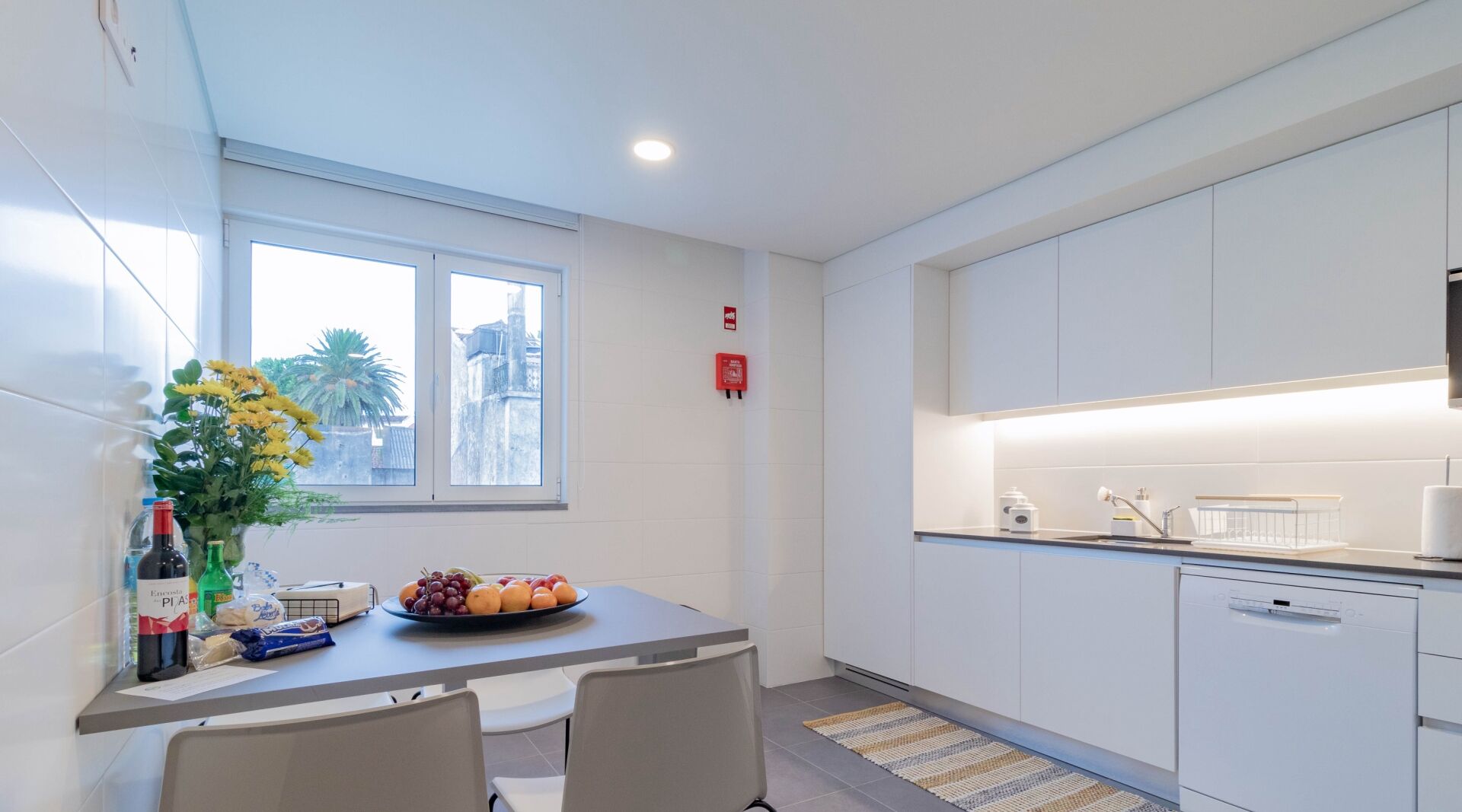 A modern kitchen with white cabinets, a dining table with chairs, a fruit bowl, and a vase of flowers.