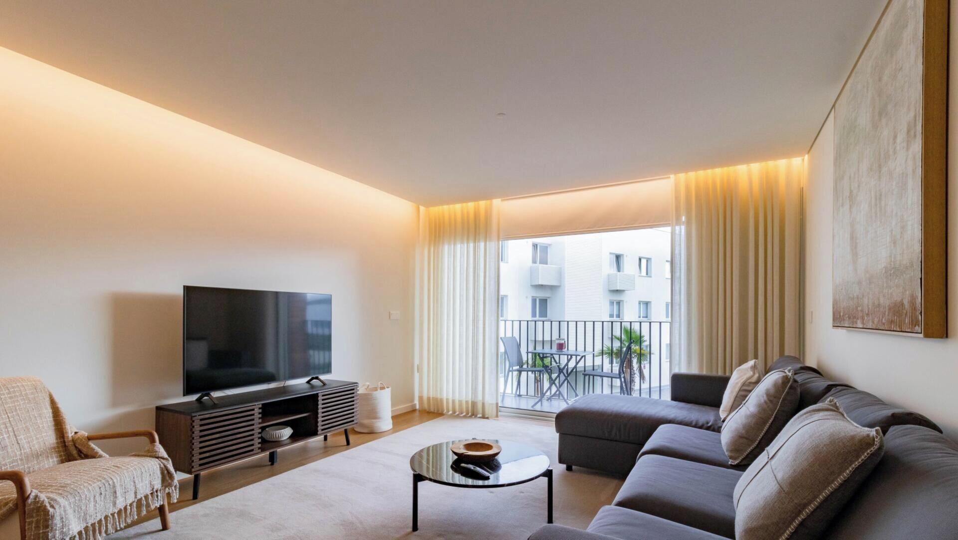 A modern living room with a flat-screen TV, a sofa, a coffee table, and a balcony with a view of buildings.