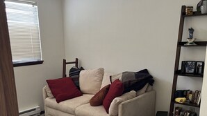 Una sala de estar con un sofá beige, cojines rojo y marrón, una mesita auxiliar negra y una estantería con diversos objetos.