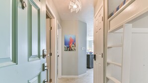 A hallway with a door on the left, a painting on the wall, and a shelving unit on the right. - New Labor Day Reduction! Sunny Beach Days at Sunrise Breeze (Galveston)