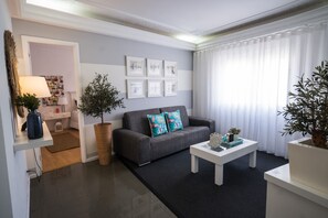 A modern living room with a grey sofa, a white coffee table, and a large window with white curtains. There are framed pictures on the wall and a potted plant on the floor. - Casa "Vó Minda" - Nazaré, 300 m from the beach. Excellent location. (Nazaré)