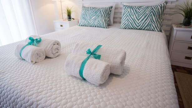 Een net opgemaakt bed met twee opgerolde handdoeken vastgebonden met een groene lint, een wit hoofdeinde en bijpassende groene en witte kussens.