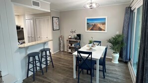 A modern kitchen with white cabinets, a bar area with stools, a dining table with chairs, and a large window with curtains. - Sandpiper Cove 1044 Beach Services  (Destin)